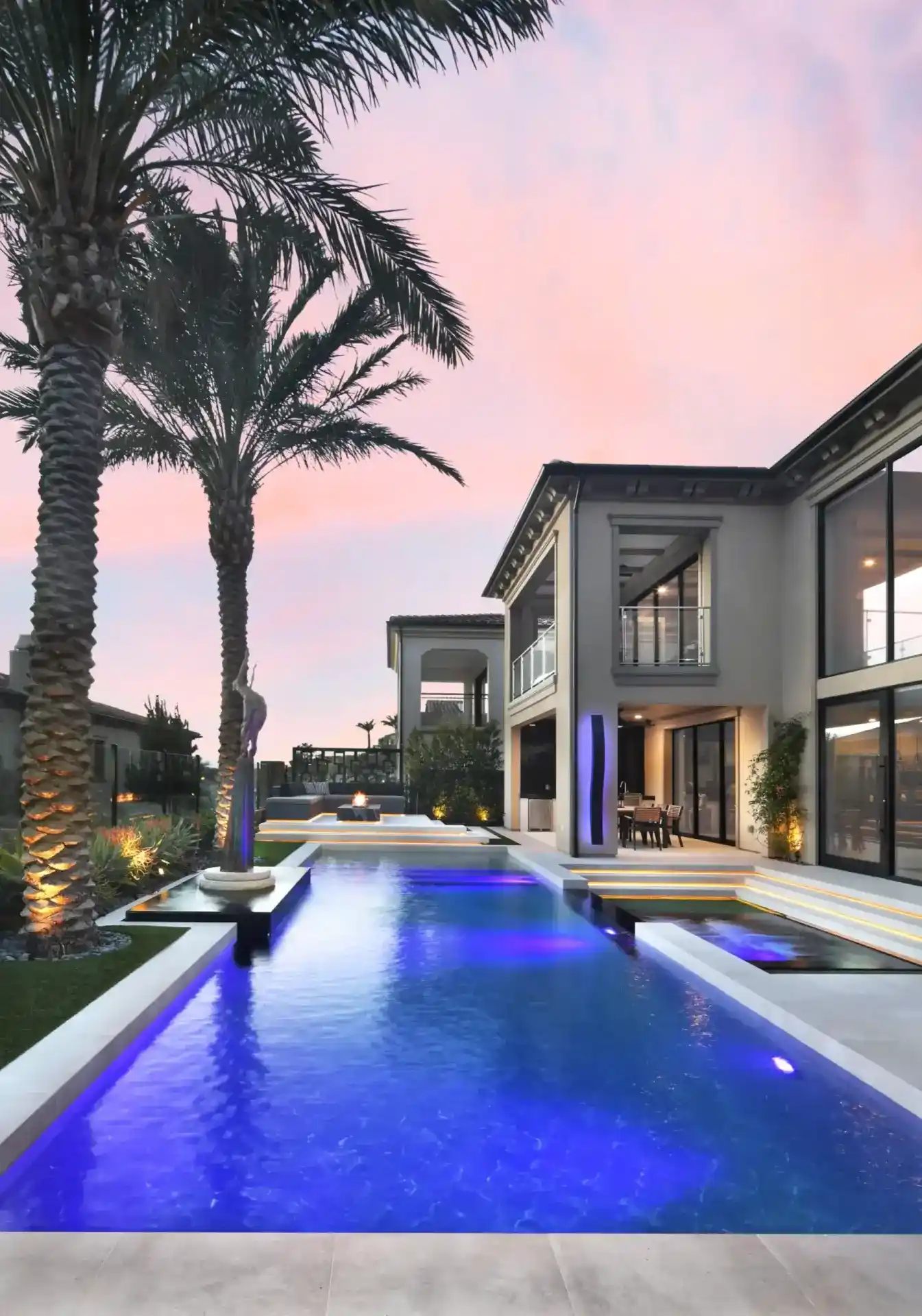 Pool lit blue at dusk beside a modern home, framed by tall palms and landscape lighting