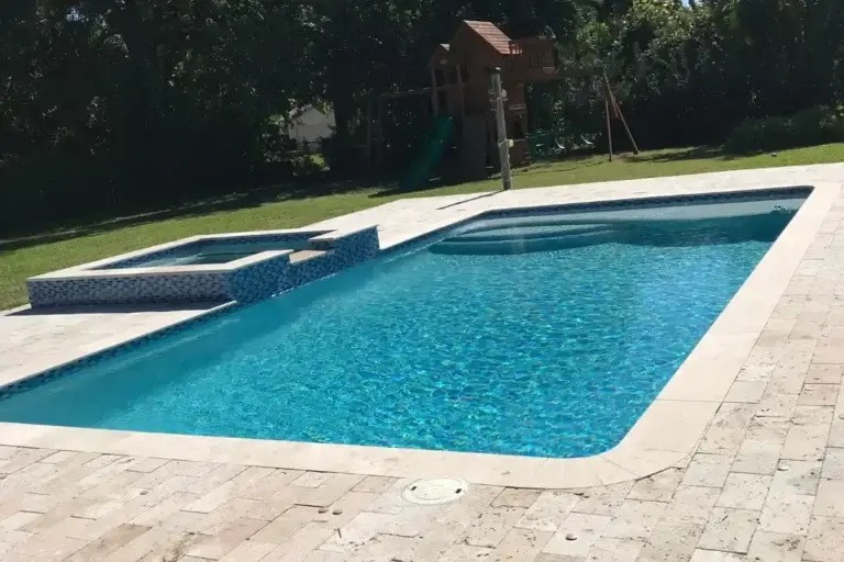 Rectangular pool with a raised spa spillway, travertine coping and a play structure beyond